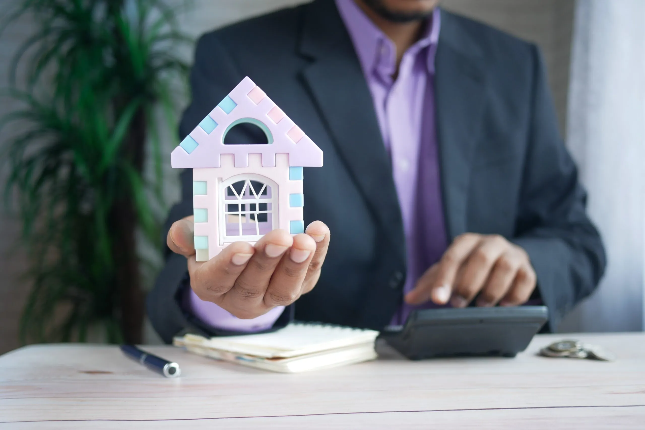 A man holding a model house in one hand while using a calculator