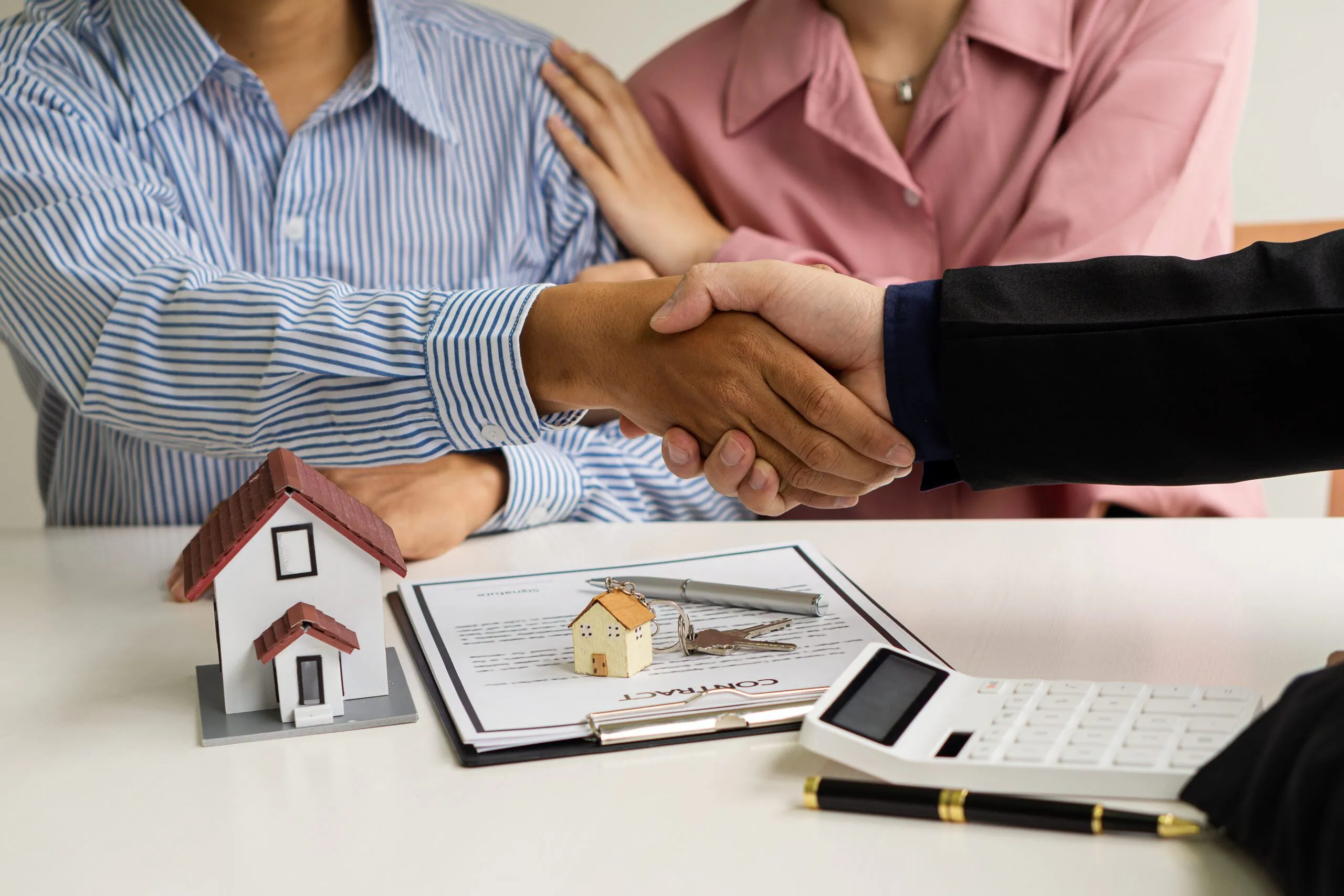 A couple shaking hands with an adviser over a model house, documents and a calculator